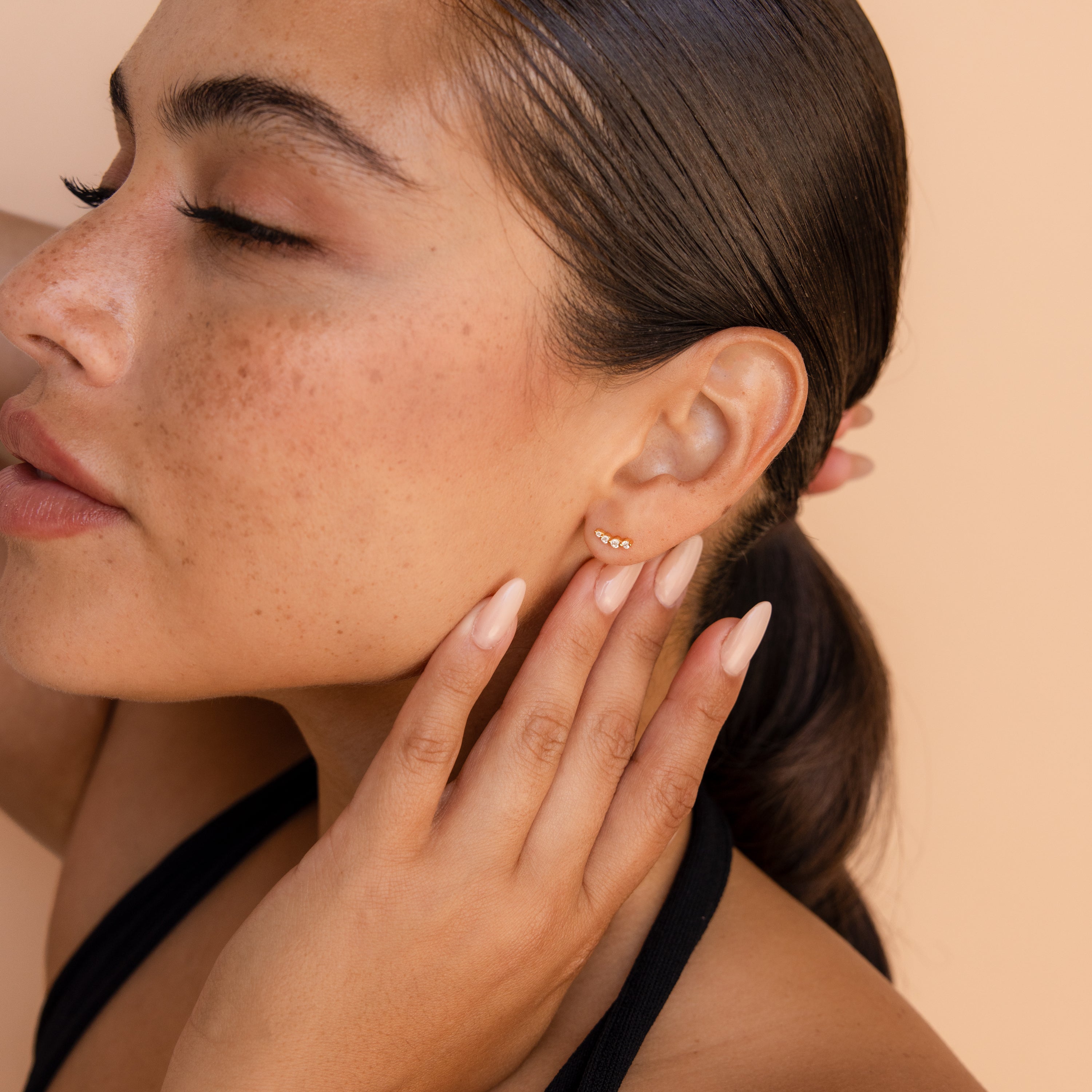 A close-up lifestyle shot of a woman with her hair pulled back, wearing a our diamond climber earring. She is touching her cheek with her hand, set against a solid warm beige background.