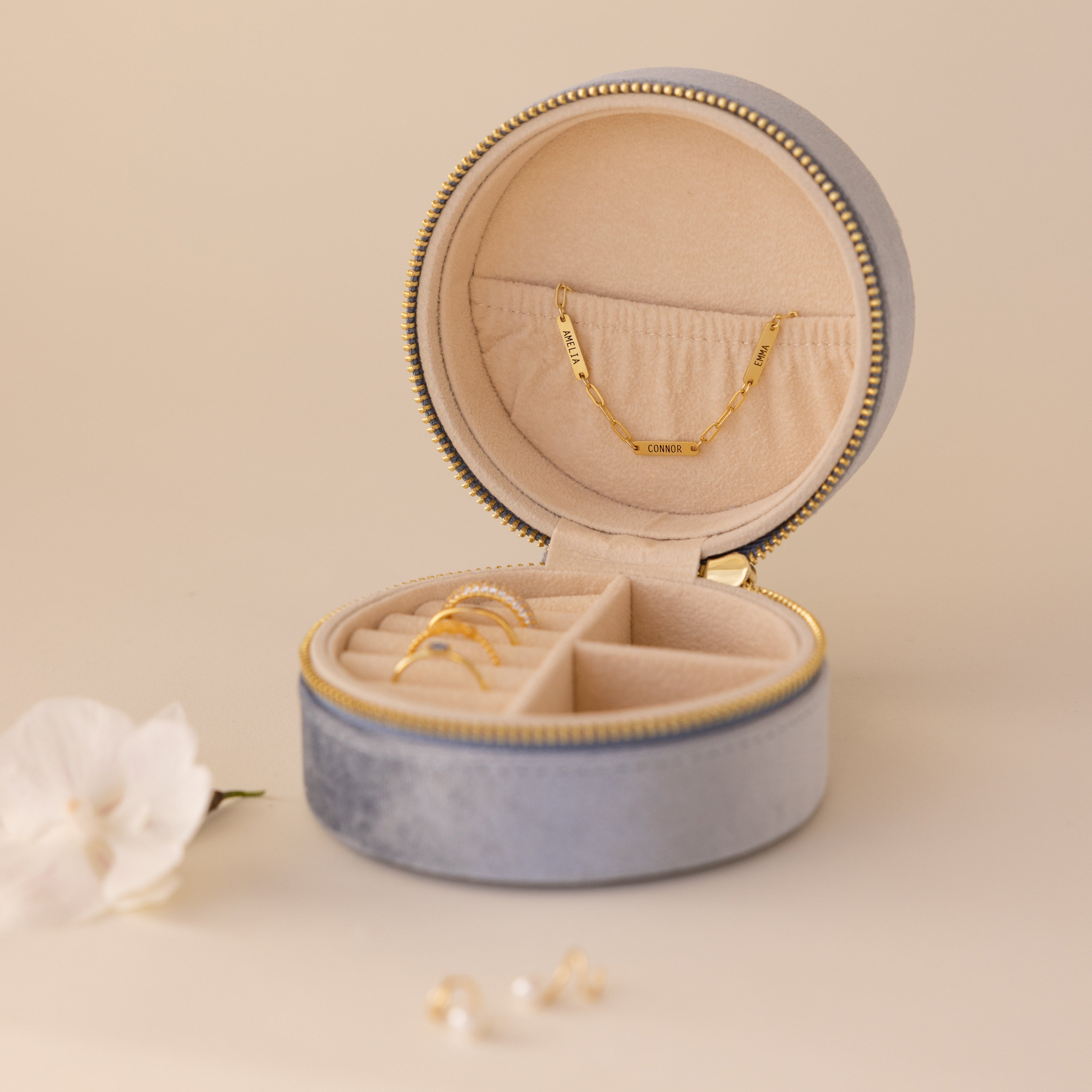 The Round Velvet Jewelry Case features gold rings and a bracelet nestled inside, placed next to a white flower on a beige surface—an elegant minimalist box that's perfect as a travel jewelry organizer.
