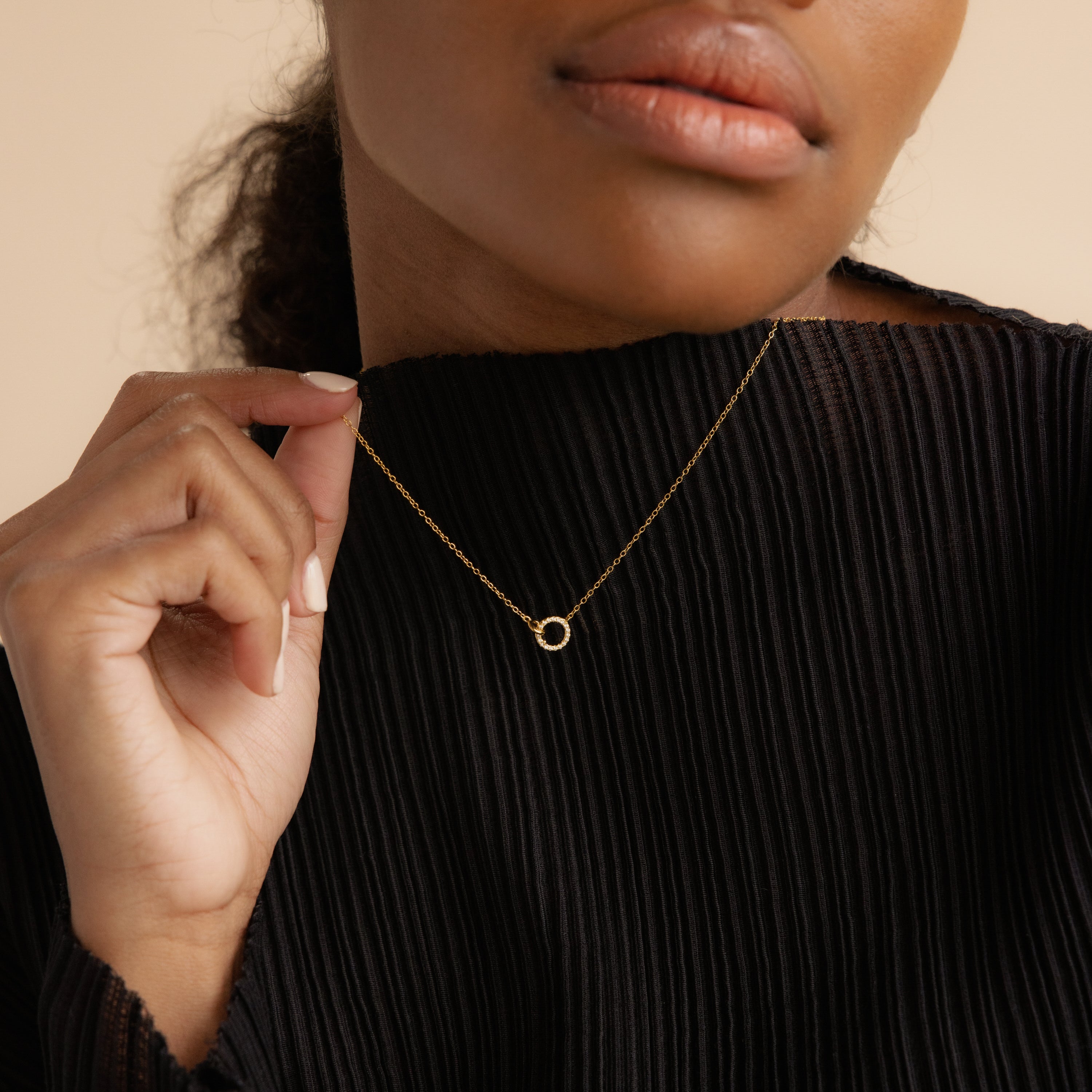 Woman in a black pleated top holds the Pave Interlocking Necklace, featuring a delicate gold design with an interlocking circular pendant—perfect for elegant unity jewelry.