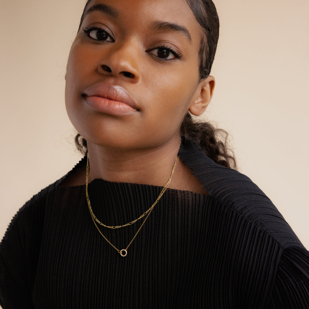 Woman with dark hair in a ponytail, wearing a black pleated top and layered gold necklaces, including the Pave Interlocking Necklace in 18K Gold, looking at the camera.