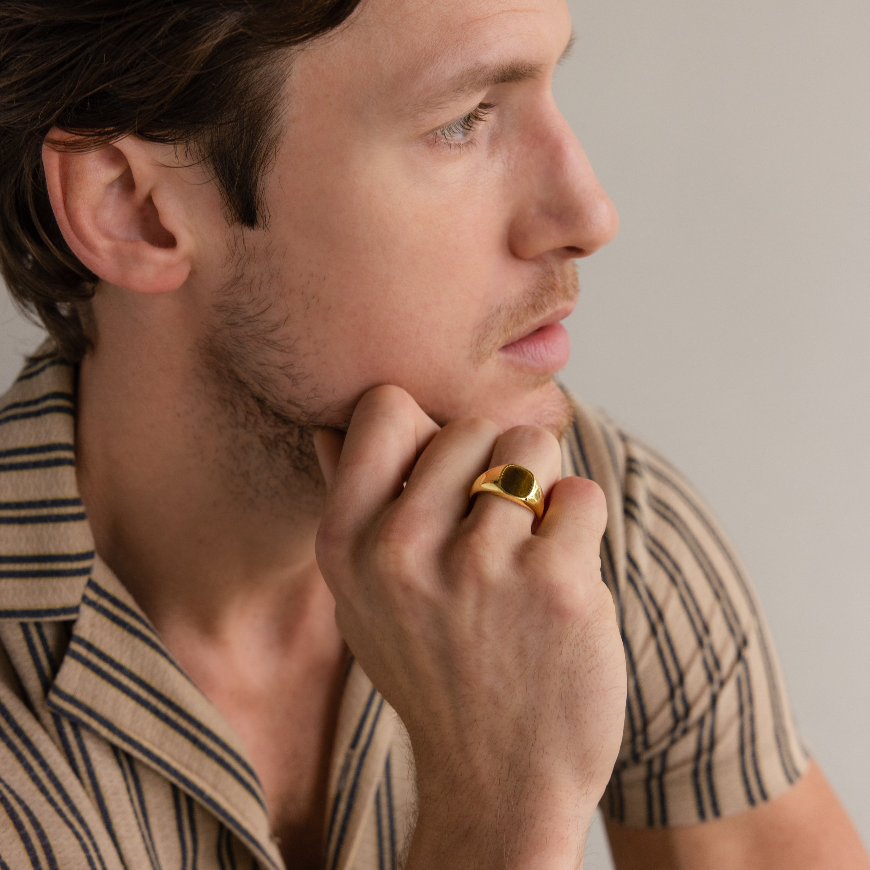 Man in striped shirt with hand on chin, wearing the Tigers Eye Signet Ring in 18K Gold, gazes thoughtfully to the side.