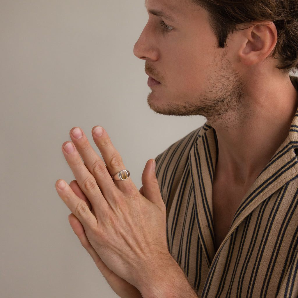 A man in a striped shirt, hands together, wearing the Tigers Eye Signet Ring in Sterling Silver, looks to the side against a plain background.
