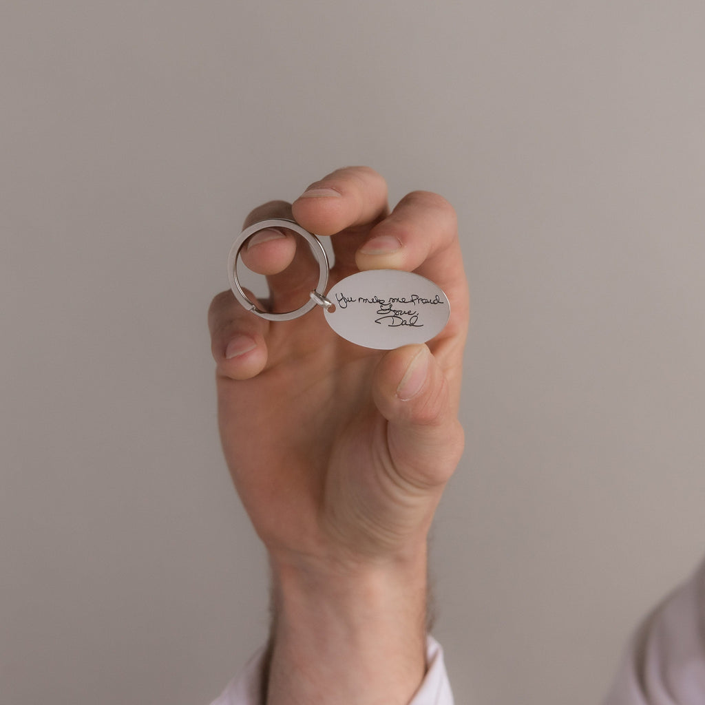 A hand holds the Handwriting Oval Keychain, featuring a small tag engraved with "You make me proud. Love, Dad.