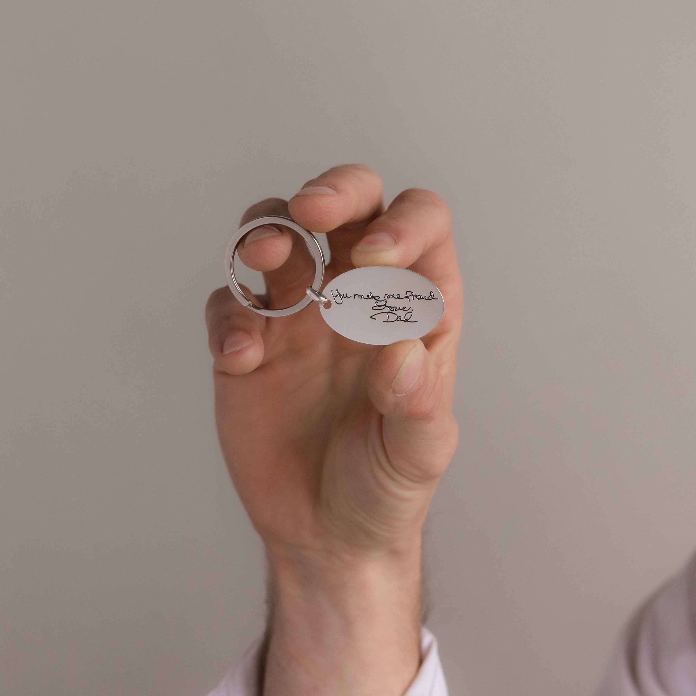 A hand holds the Handwriting Oval Keychain, featuring a small tag engraved with "You make me proud. Love, Dad.