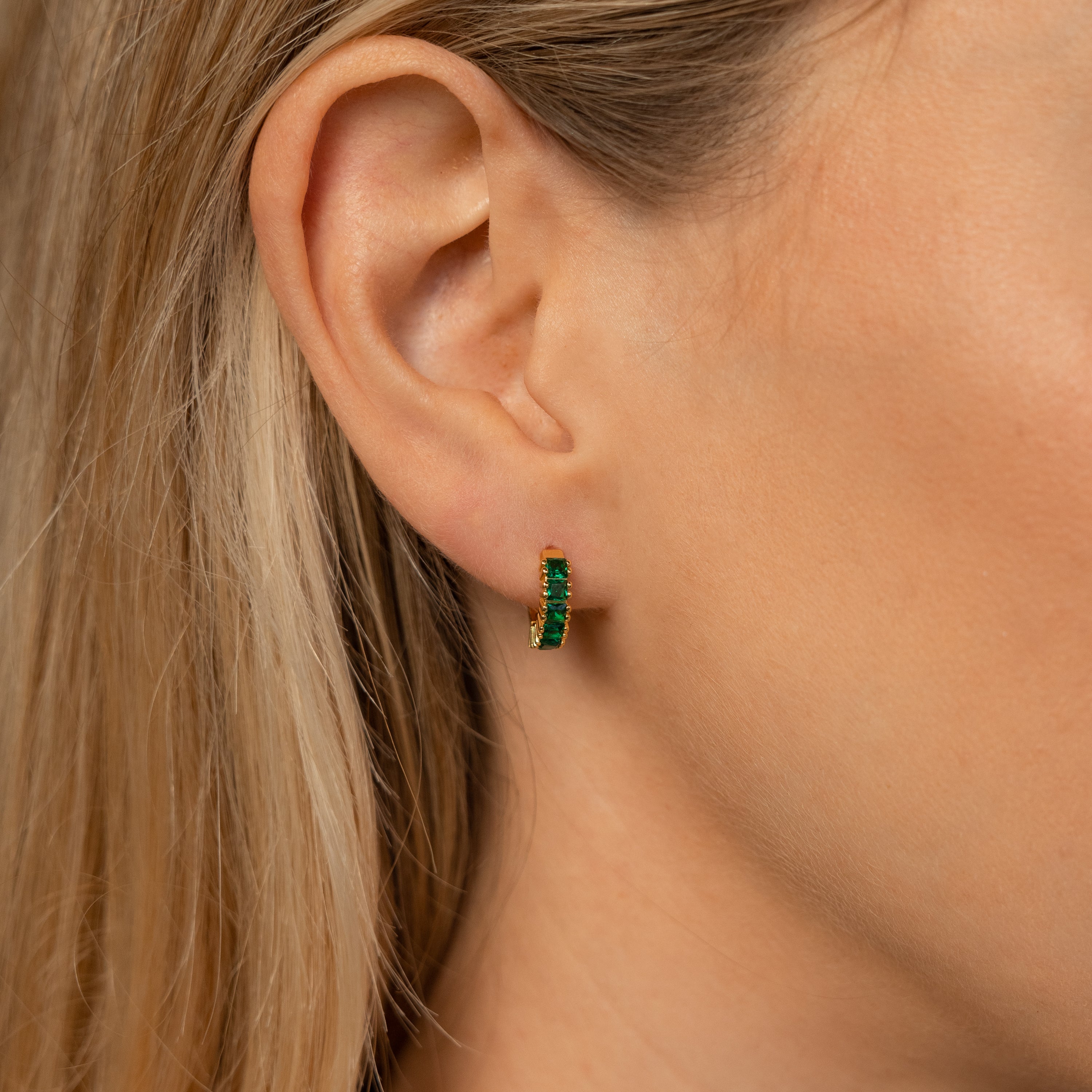 Close-up of a woman's ear wearing Genevieve Emerald Huggies—small gold hoops with green gemstones—perfect elegant birthstone earrings.