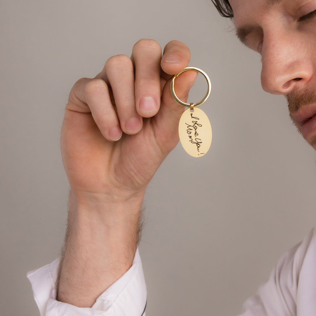 A person holding the Handwriting Oval Keychain with a gold tag engraved in elegant cursive that says "I love you, Mom.