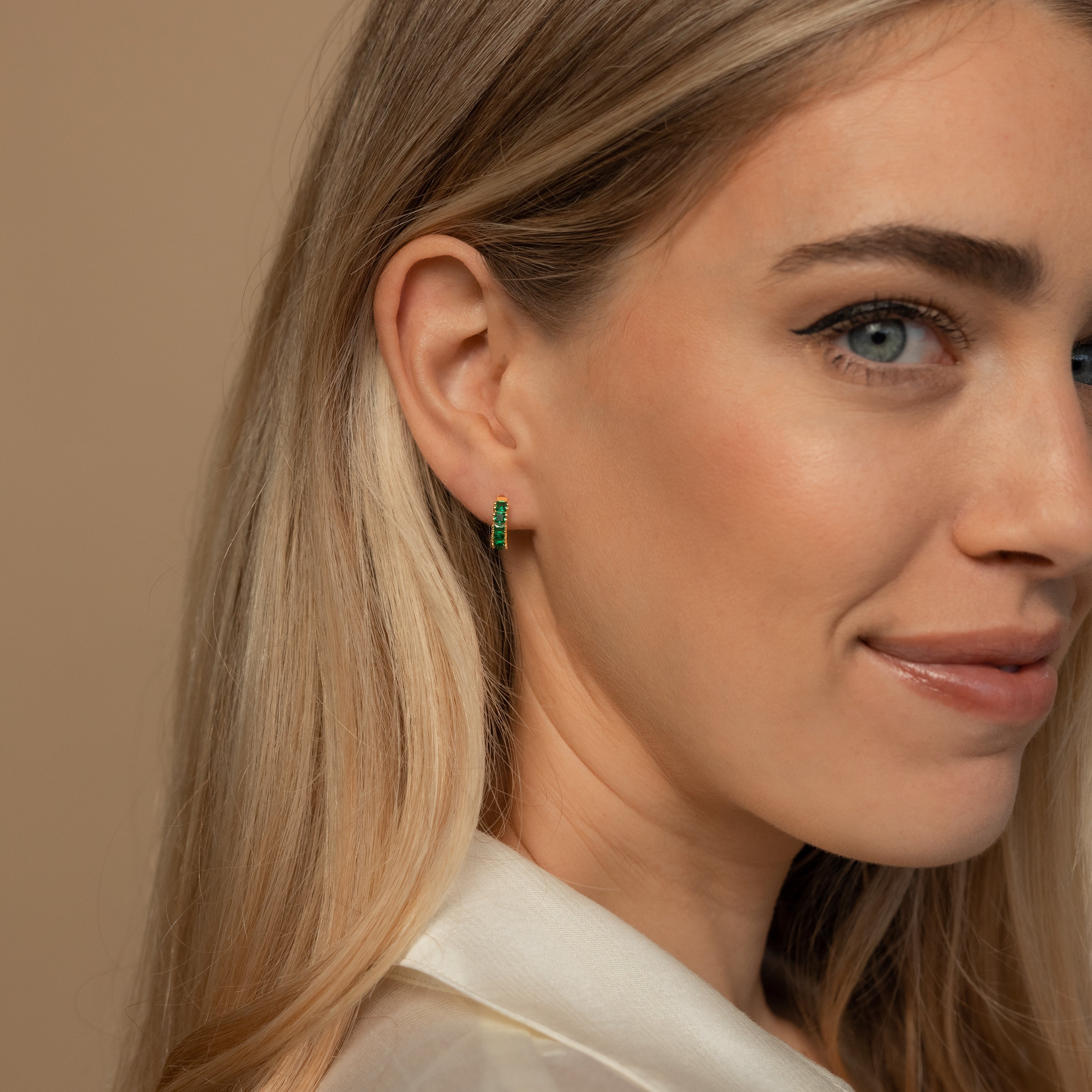 Woman with long blonde hair wears the Genevieve Emerald Huggies—small green earrings—while smiling slightly against a beige background.