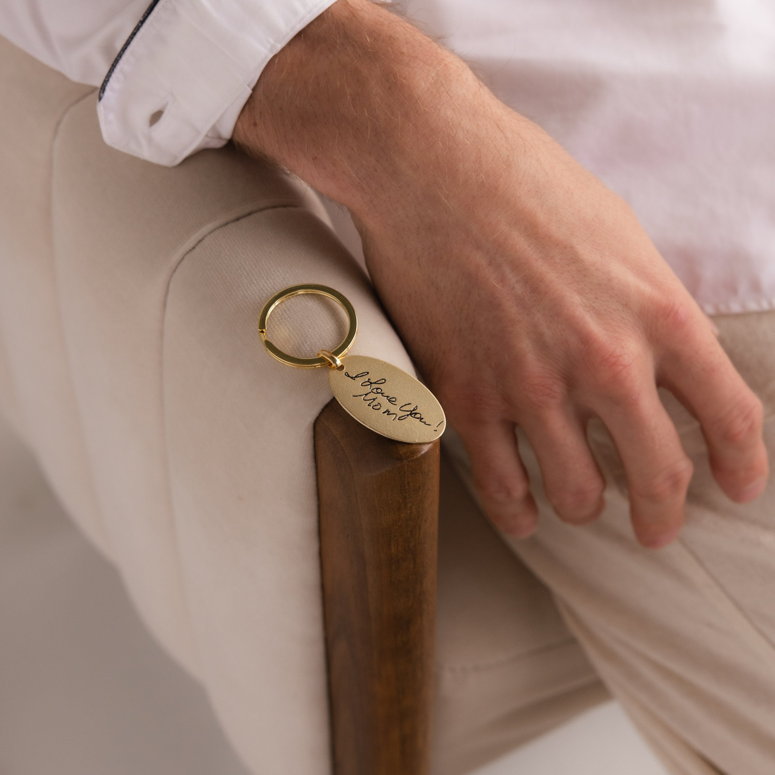 A hand rests on a chair arm, holding the Handwriting Oval Keychain engraved with elegant cursive writing.