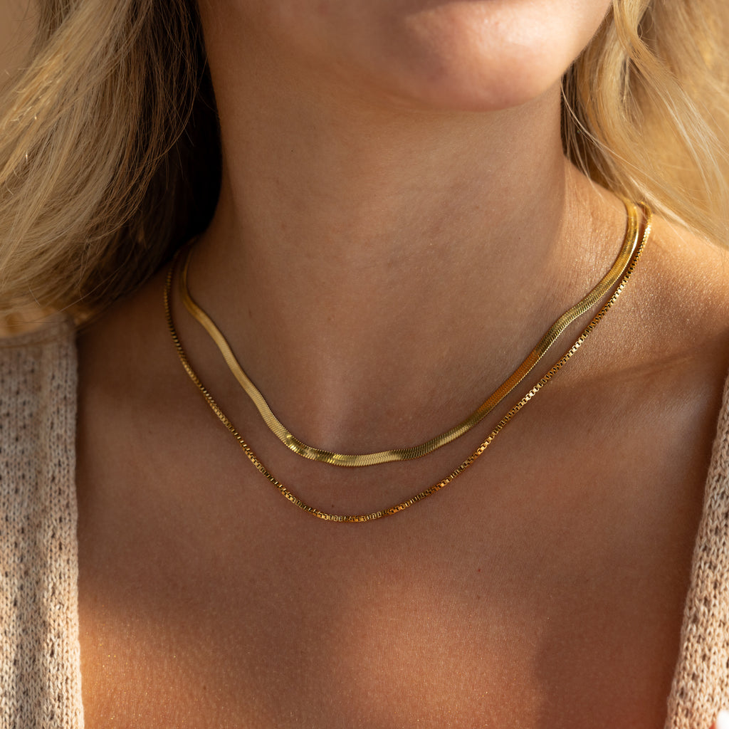 Close-up of a woman wearing the Herringbone Duo Chain Necklace in 18K Gold layered with a box chain, styled with a textured beige top and blonde hair.