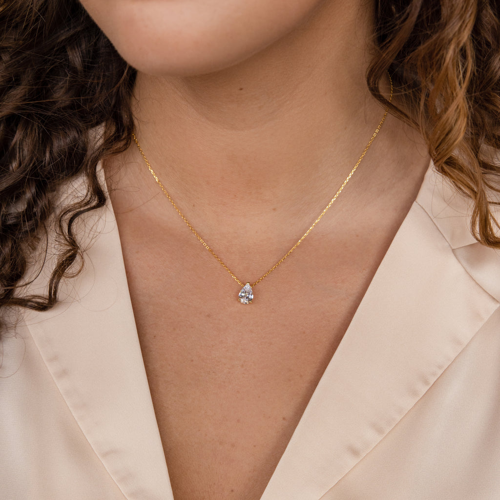 A woman wears the Ophelia Birthstone Necklace, featuring a minimalist gold teardrop pendant, over a cream-colored blouse.