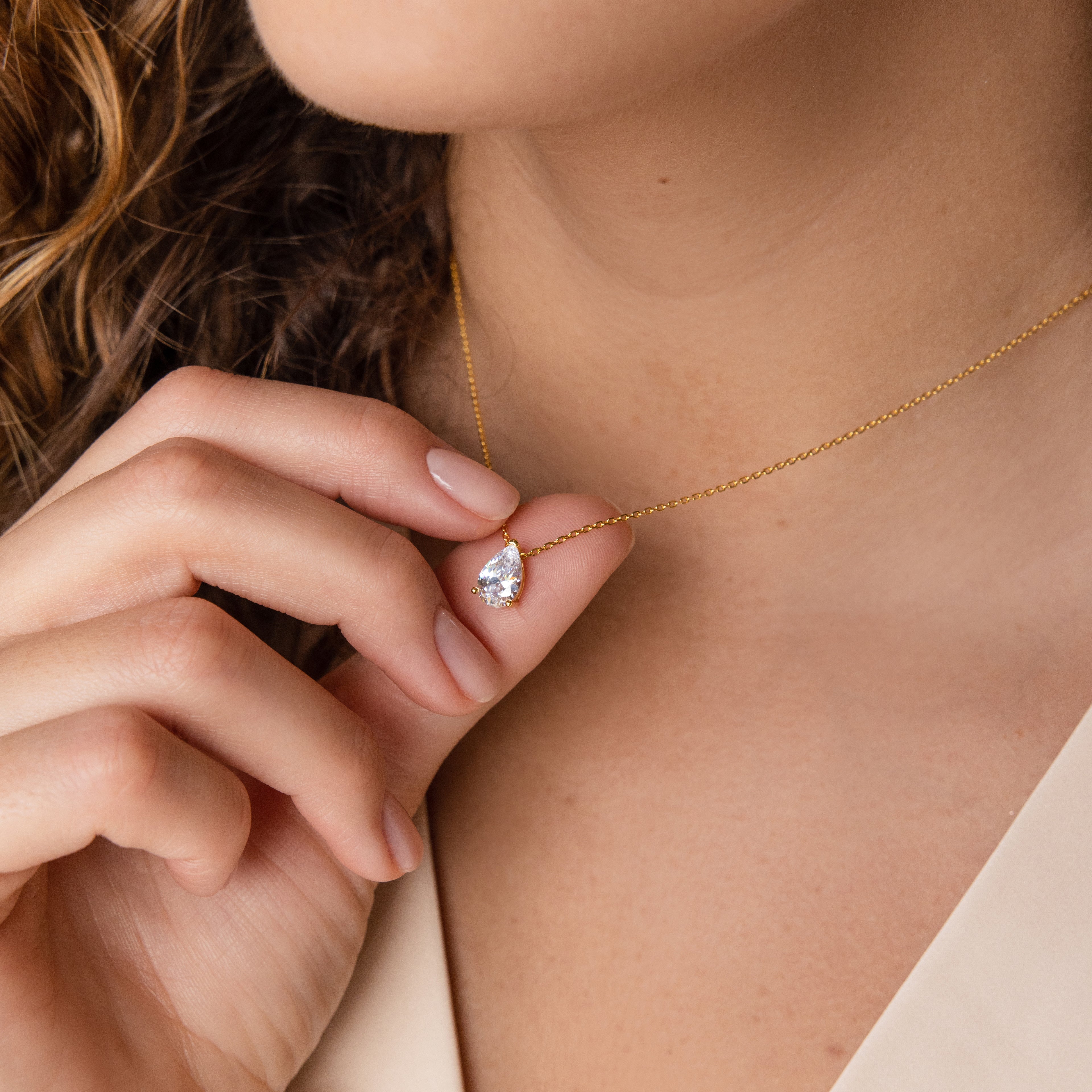 A woman holds the Ophelia Topaz Necklace, featuring a gold teardrop-shaped pendant, close to her neck.