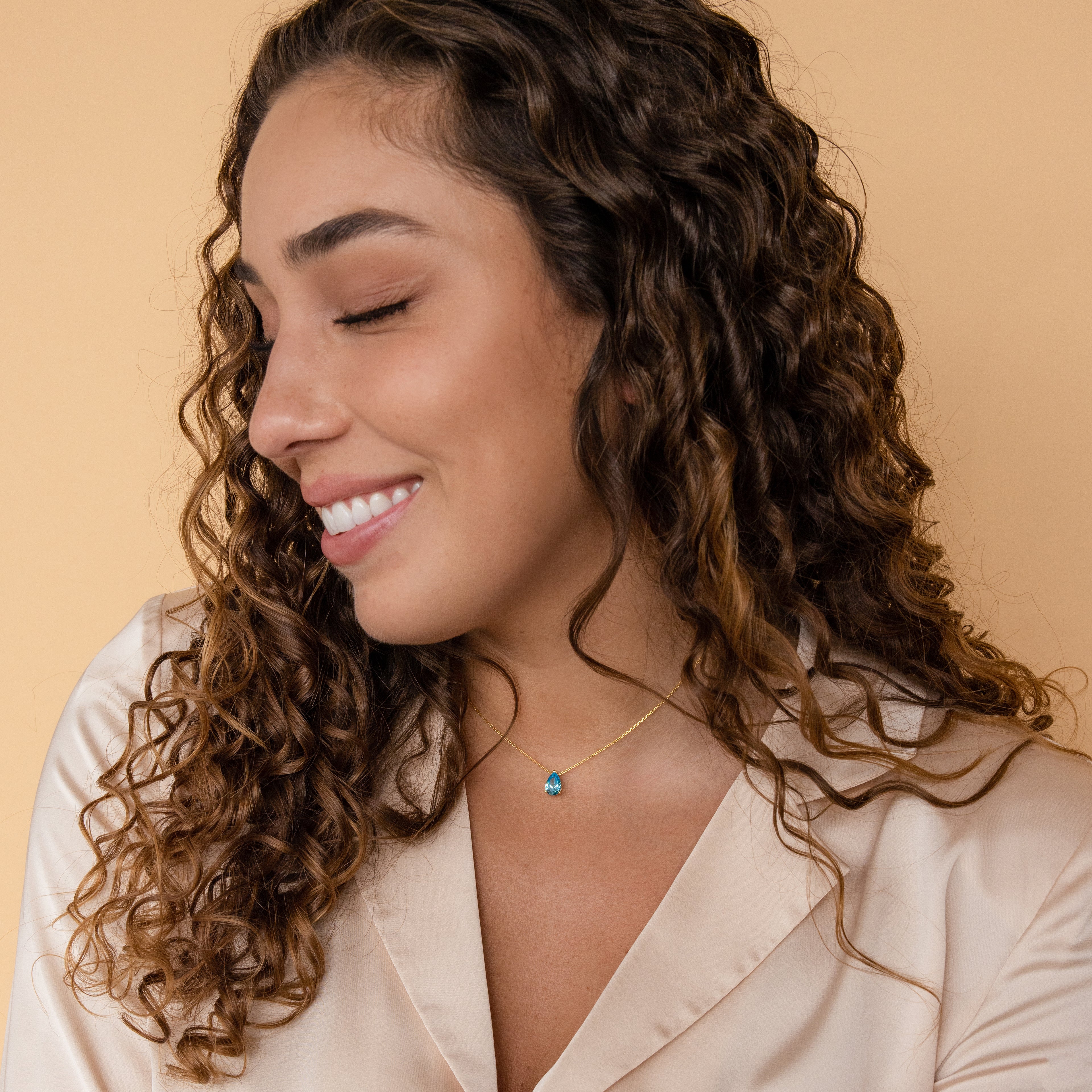 A woman with curly hair smiles with eyes closed, wearing a cream blouse and the Ophelia Birthstone Necklace featuring a delicate gold chain and blue pendant.