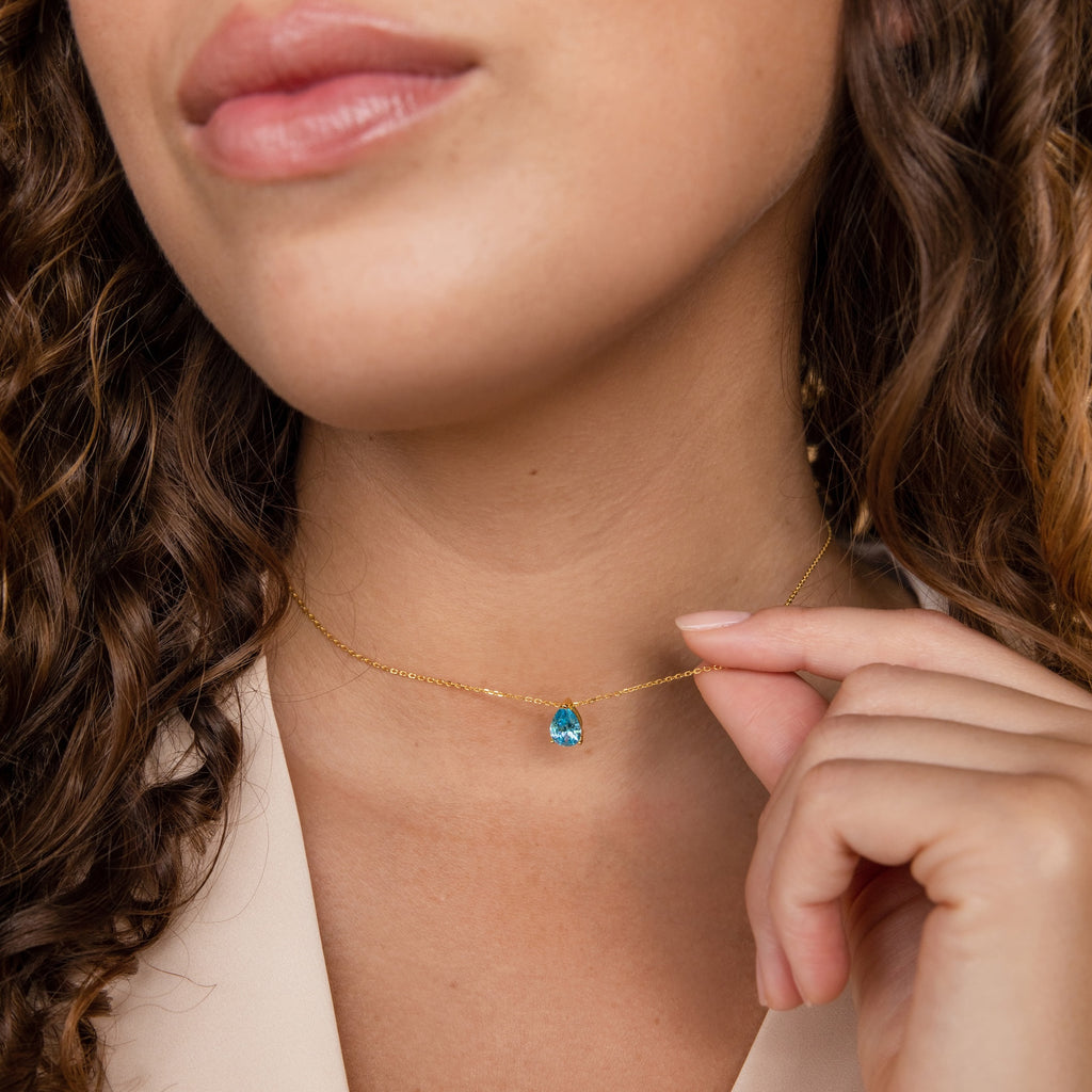 A woman with curly hair delicately holds the Ophelia Birthstone Necklace, a minimalist gold piece featuring a blue gemstone pendant.