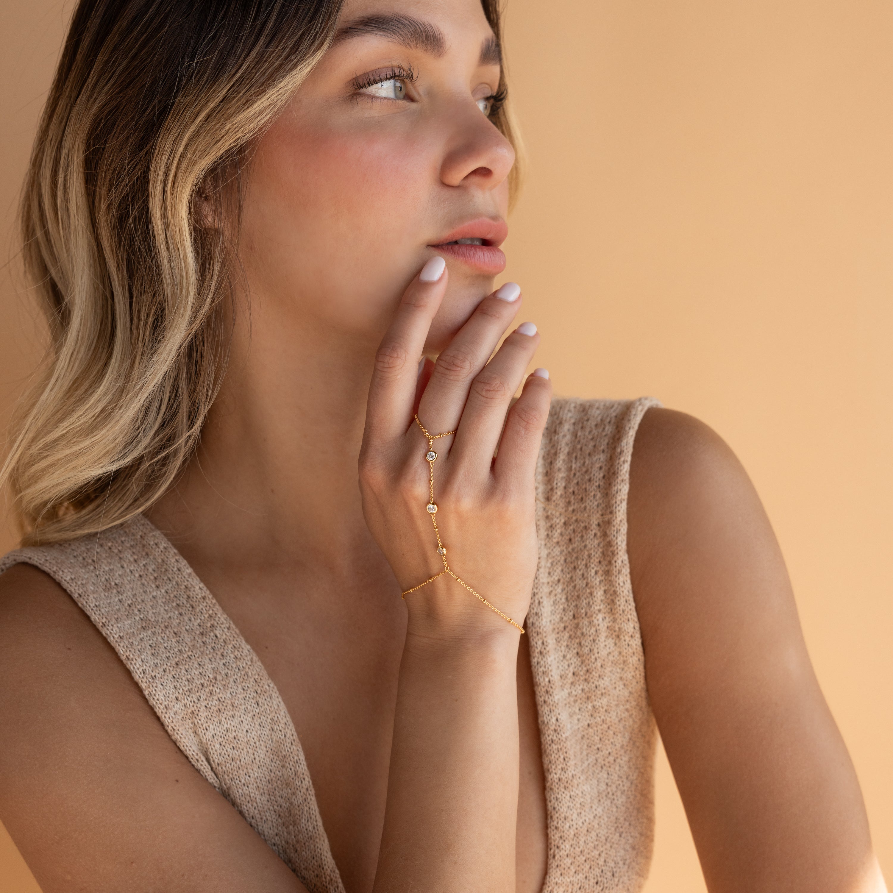 Woman with wavy hair wears the Diamond Bezel Hand Chain in 18K Gold and a sleeveless beige top, gazing to the side against a tan background.