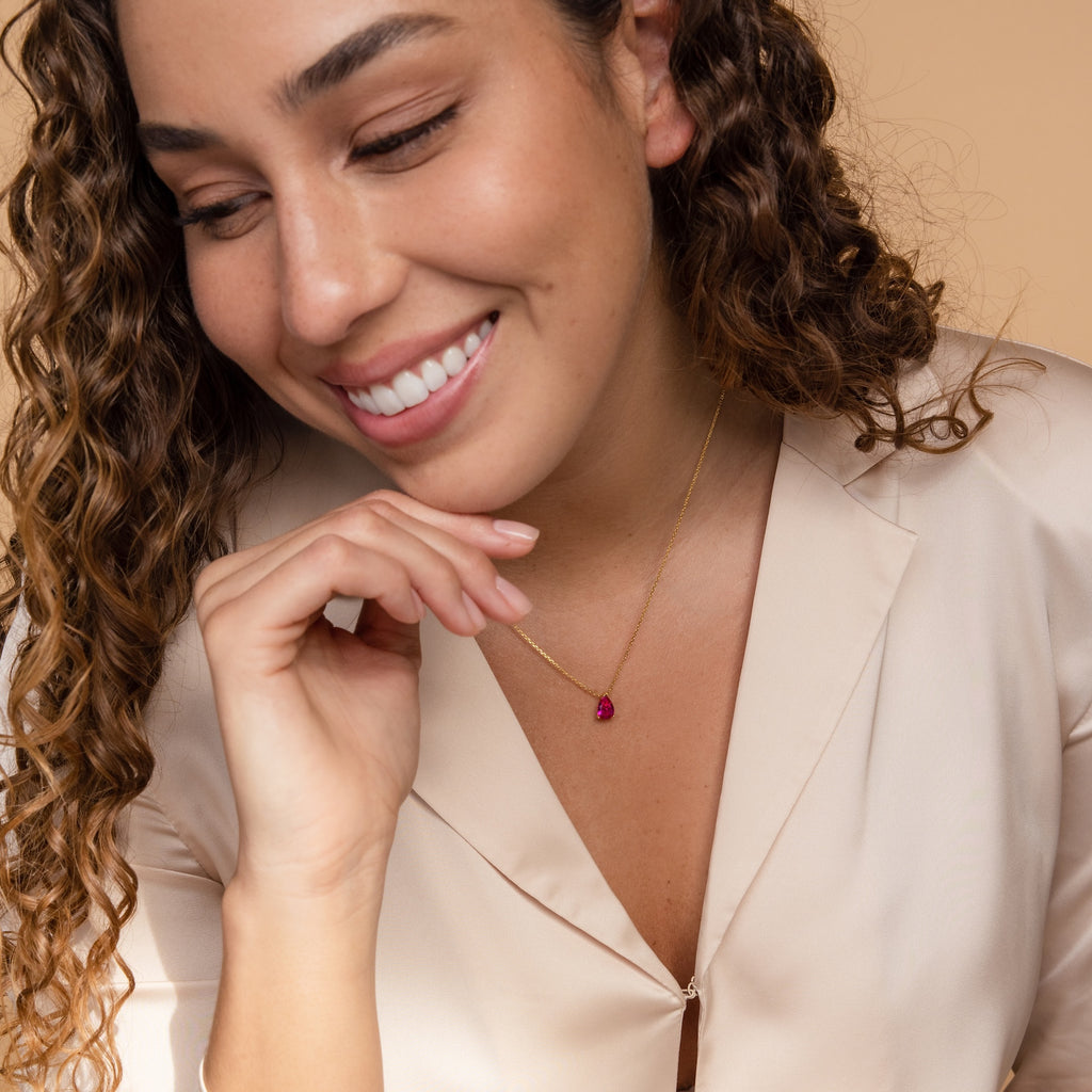 Smiling woman with curly hair wearing a beige blouse and the Ophelia Birthstone Necklace featuring a gold chain and red pendant.