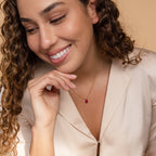 Smiling woman with curly hair wearing a beige blouse and the Ophelia Birthstone Necklace featuring a gold chain and red pendant.