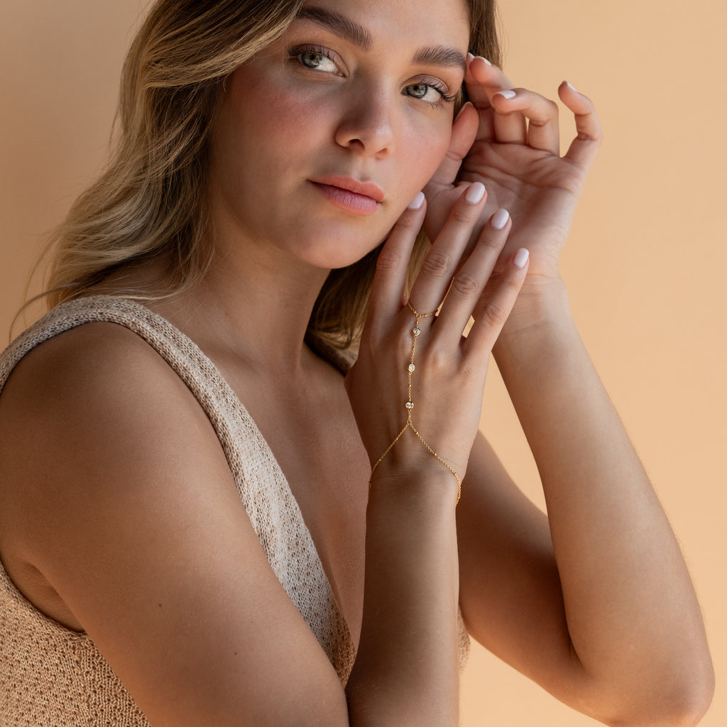 A woman with light brown hair wearing a knitted top and the Diamond Bezel Hand Chain poses against a beige background.