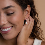 A close-up of a smiling woman with curly brown hair wearing a single silver Dangling Pearl Drop Earring. The earring features a polished silver hook with an oval freshwater pearl suspended at the center. She gently touches her ear with one hand, showcasing the delicate charm of the earring.