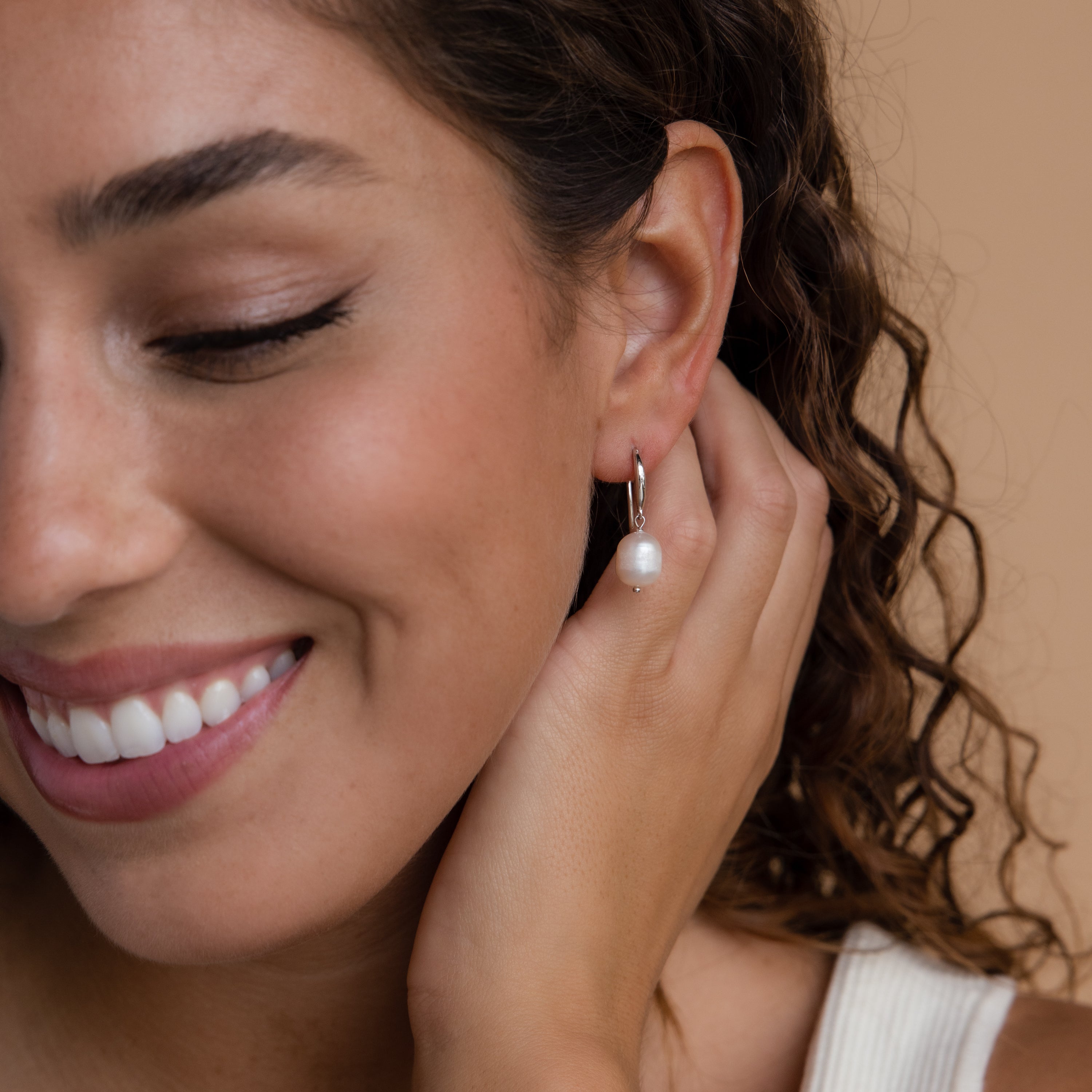A close-up of a smiling woman with curly brown hair wearing a single silver Dangling Pearl Drop Earring. The earring features a polished silver hook with an oval freshwater pearl suspended at the center. She gently touches her ear with one hand, showcasing the delicate charm of the earring.