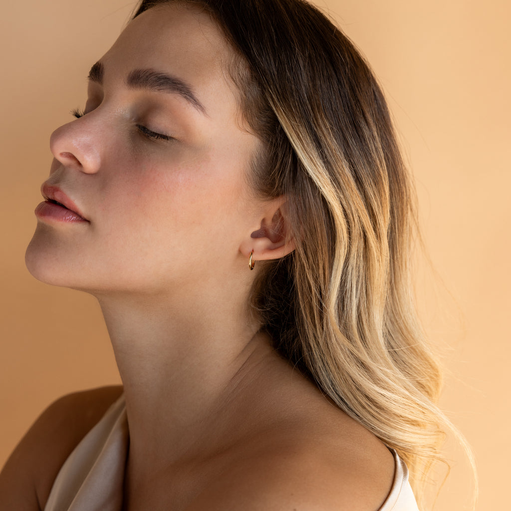 A woman with closed eyes and wavy blonde hair poses against a soft beige background, wearing the Small Bold Hoops in 18K Gold—minimalist earrings that add an elegant touch for everyday wear.