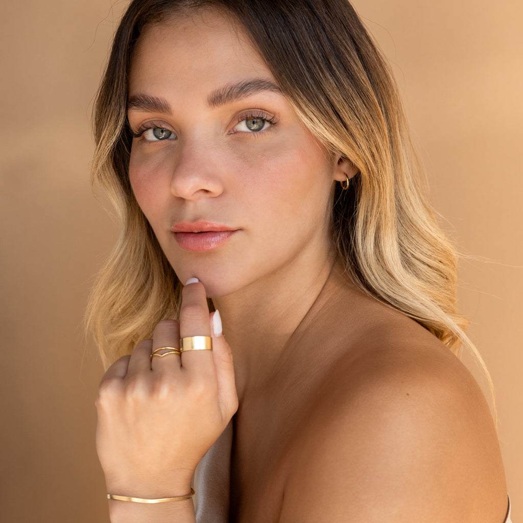 Woman with wavy blonde hair wears Small Bold Hoops in 18K Gold, posing against a beige background and looking at the camera.