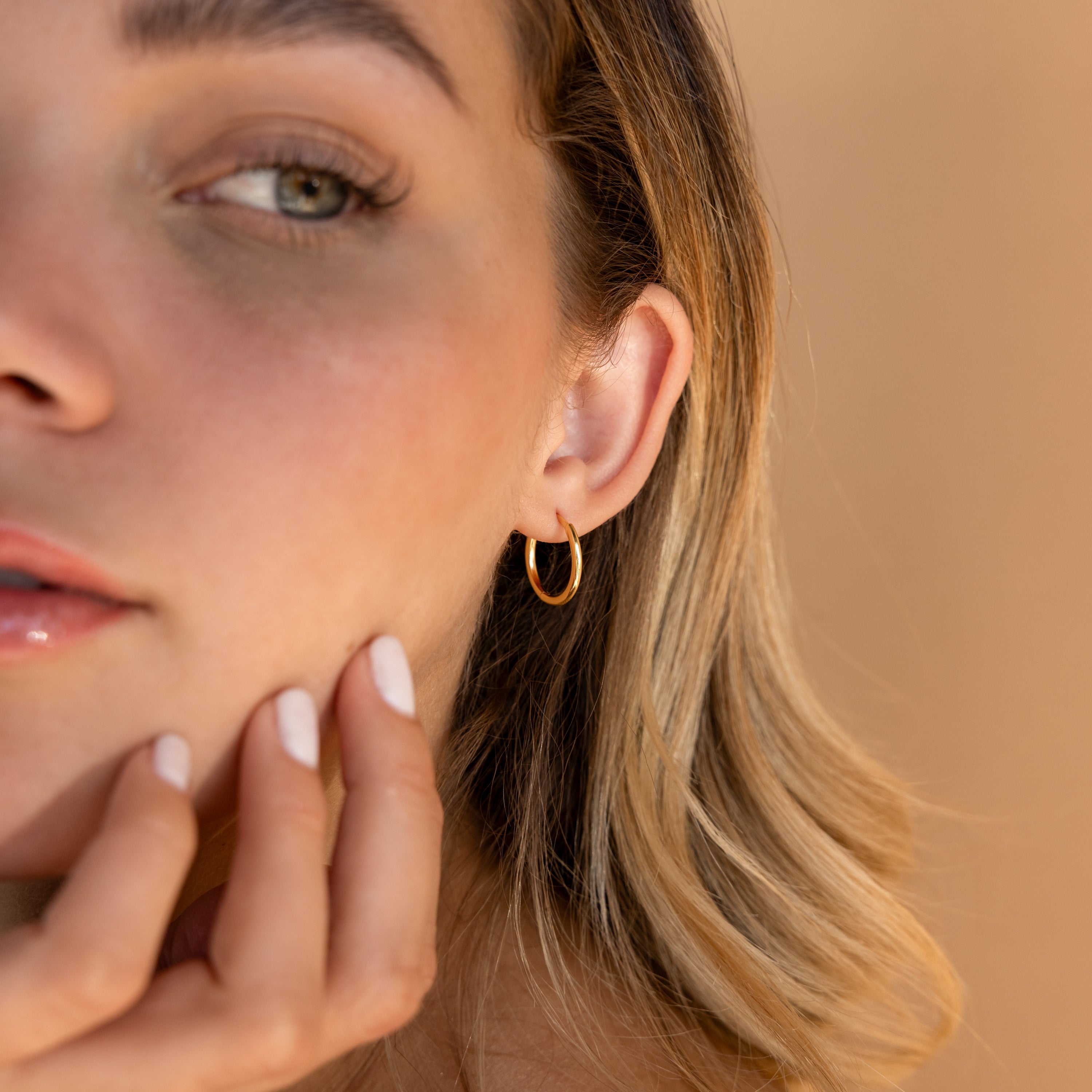 Close-up of a woman wearing the Medium Bold Hoops in 18K Gold, stylish lightweight earrings ideal for everyday wear, as she touches her chin with her hand.