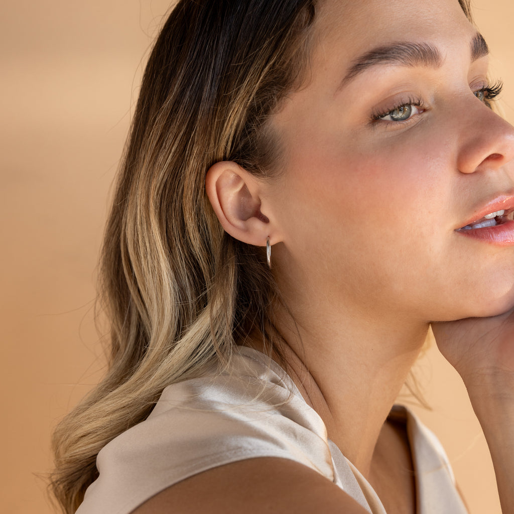 A woman with wavy blonde hair wears the Medium Bold Hoops in Sterling Silver, appearing thoughtful against a beige background and highlighting the elegance of these everyday earrings.