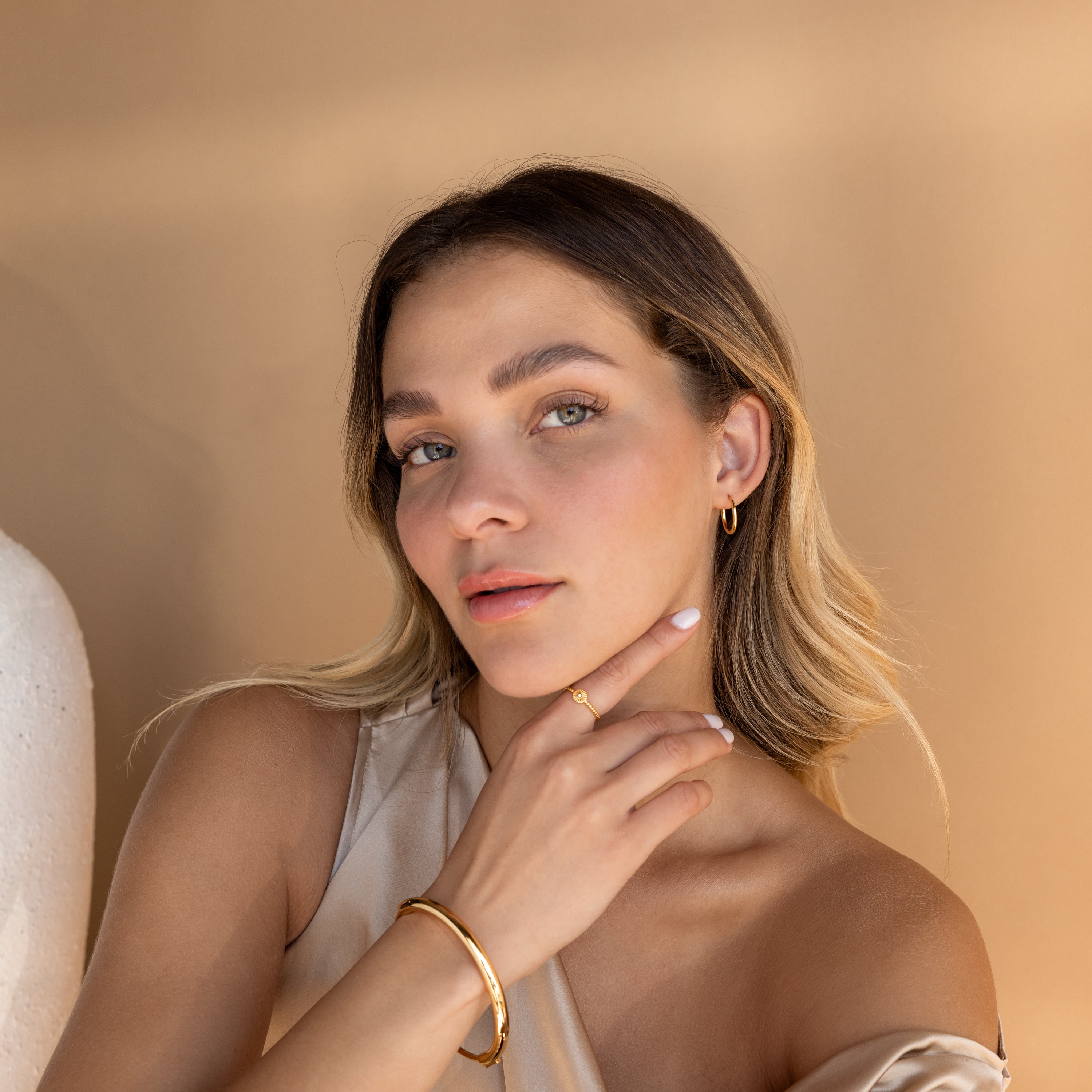 A woman with light brown hair poses against a beige background, wearing a neutral top and everyday jewelry, including Medium Bold Hoops.