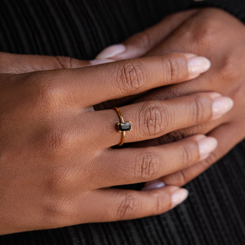 A hand wearing the Black Emerald Ring—a vintage-style gold Art Deco ring with a rectangular black stone—rests on another hand against a dark background.