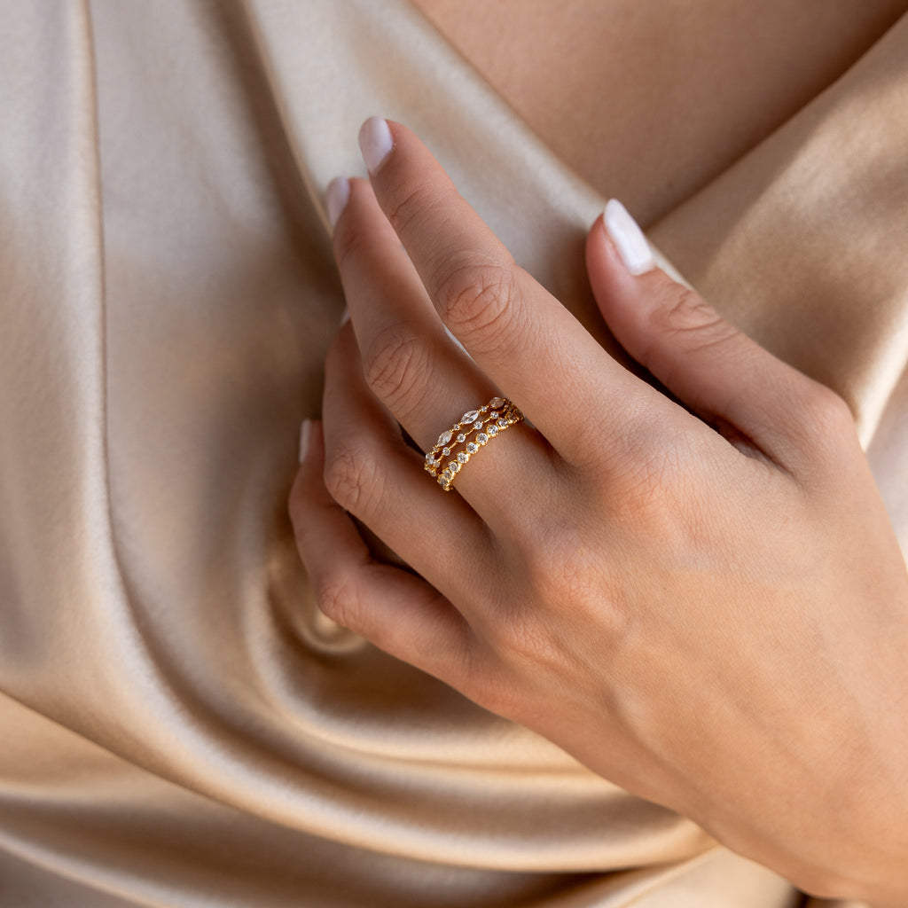 A hand with manicured nails wearing elegant jewelry, including a Crystal Stacking Ring Set, rests on champagne-colored satin fabric.