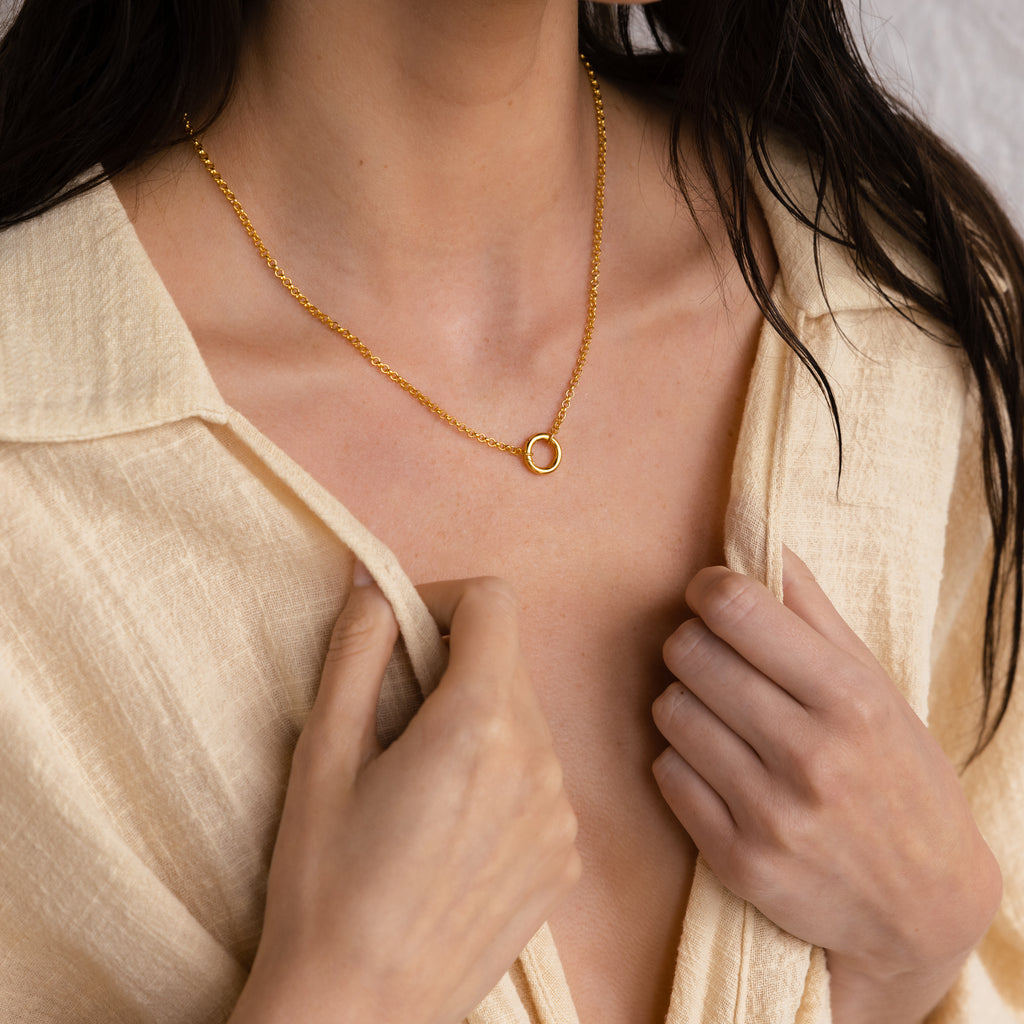 A woman in a beige shirt wears the minimalist Rolo Circle Charm Necklace—a simple gold accent—holding her collar slightly open.