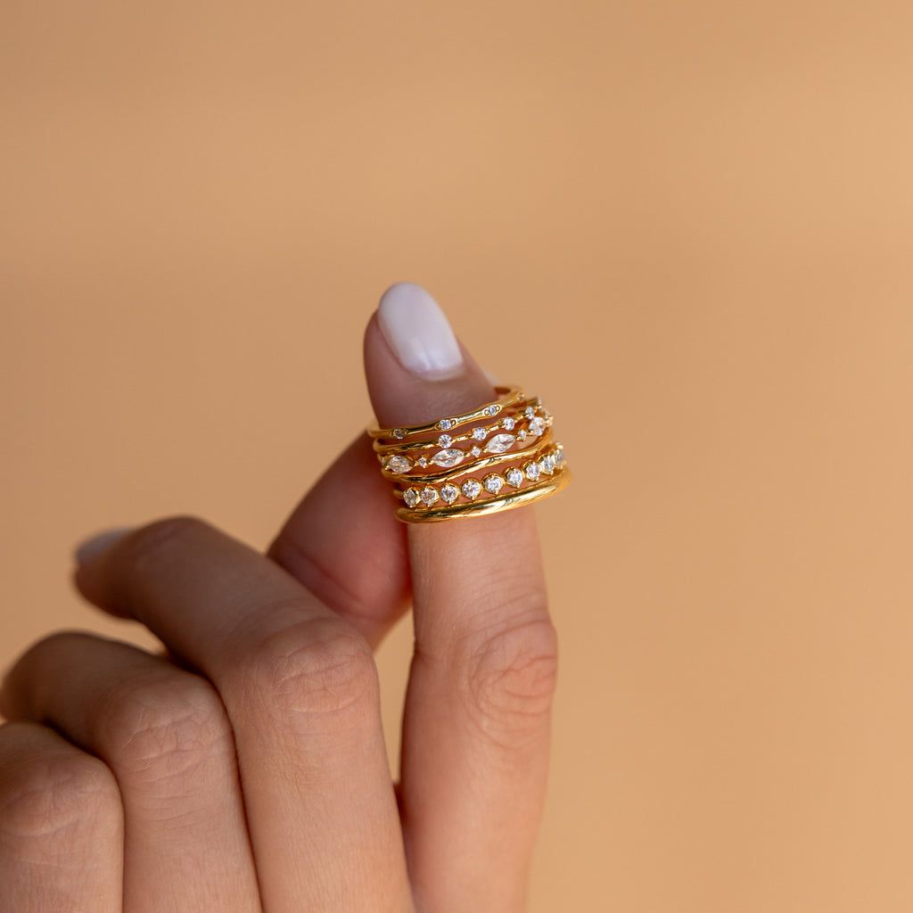 A hand with manicured nails wears the Crystal Stacking Ring Set on the index finger, displaying elegant gold and diamond-inspired rings against a beige background.