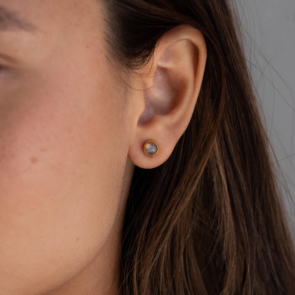 A close-up of a woman's ear wearing Moonstone Bezel Studs—these minimal earrings feature luminous moonstones in delicate, round gold settings that subtly catch the light.