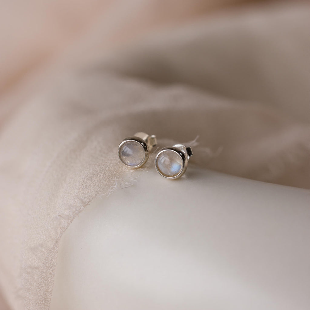 Moonstone Bezel Studs feature round, translucent gemstones set in gold, resting on soft beige fabric—ideal earrings for those who love an ethereal, understated jewelry style.