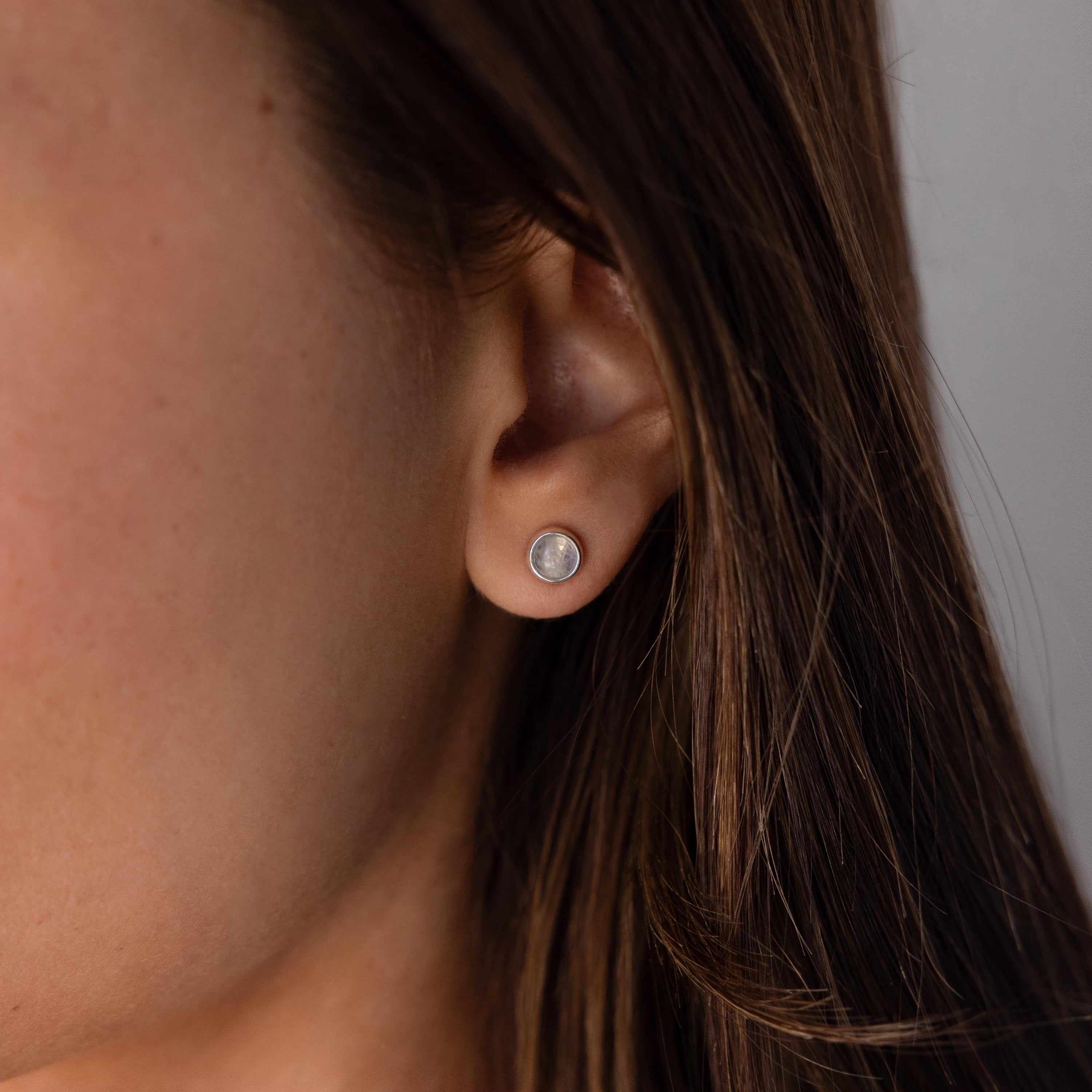 Close-up of a woman’s ear with long brown hair, wearing Moonstone Bezel Studs—delicate earrings that bring a soft, ethereal glow to any style.