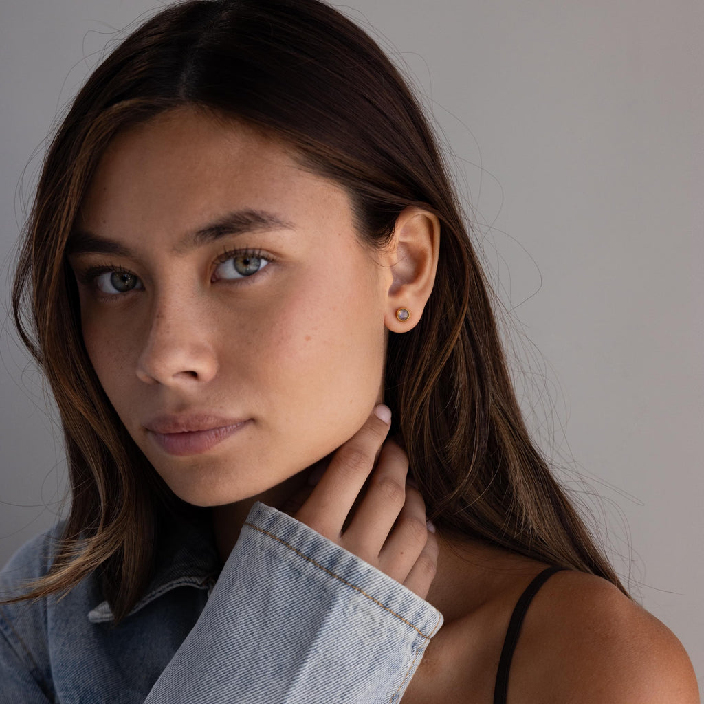 A woman with long brown hair in a denim jacket wears Moonstone Bezel Studs—understated jewelry that adds a subtle shimmer—as she looks at the camera.