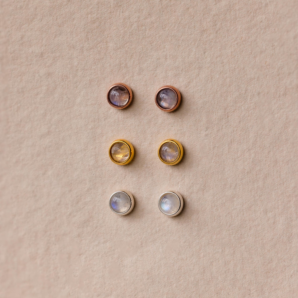 Six pairs of round stud earrings, including the Moonstone Bezel Studs, come in gold, silver, and rose gold settings for a subtle, ethereal look displayed on a beige background.