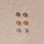 Six pairs of round stud earrings, including the Moonstone Bezel Studs, come in gold, silver, and rose gold settings for a subtle, ethereal look displayed on a beige background.