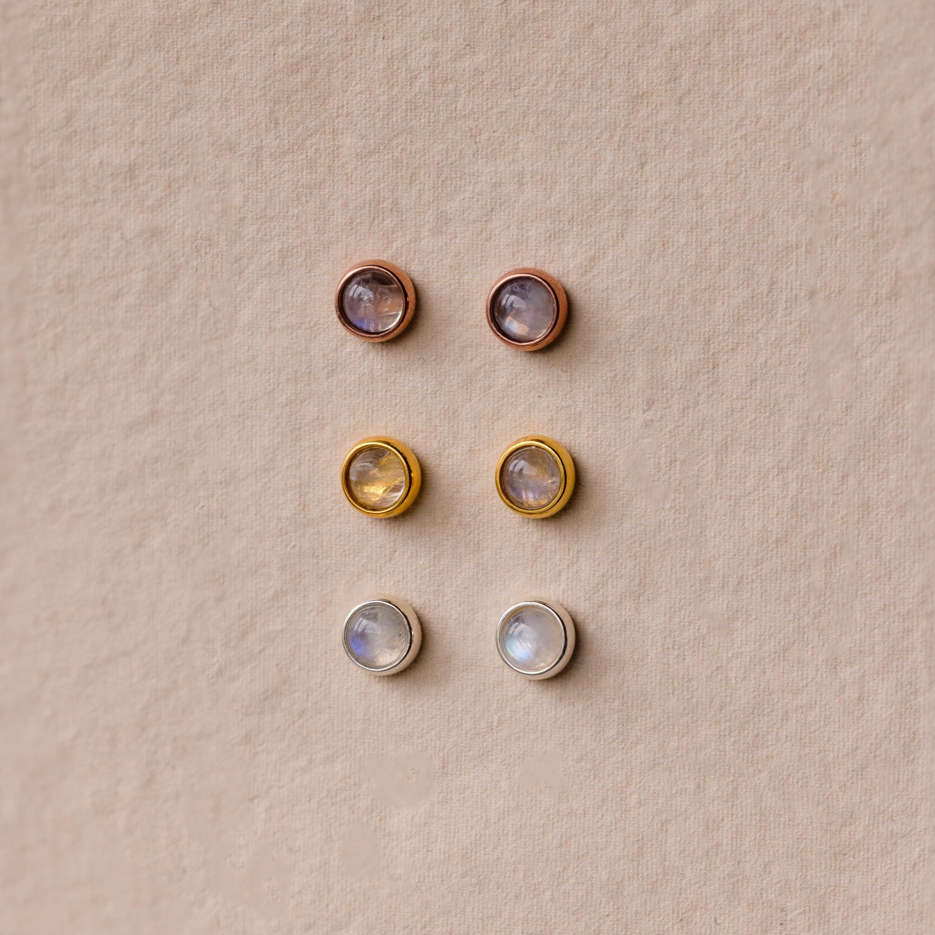 Six pairs of round stud earrings, including the Moonstone Bezel Studs, come in gold, silver, and rose gold settings for a subtle, ethereal look displayed on a beige background.