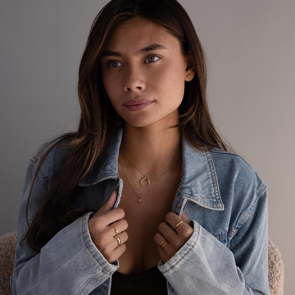 Woman in a denim jacket wearing Moonstone Bezel Studs, sitting indoors against a neutral background.