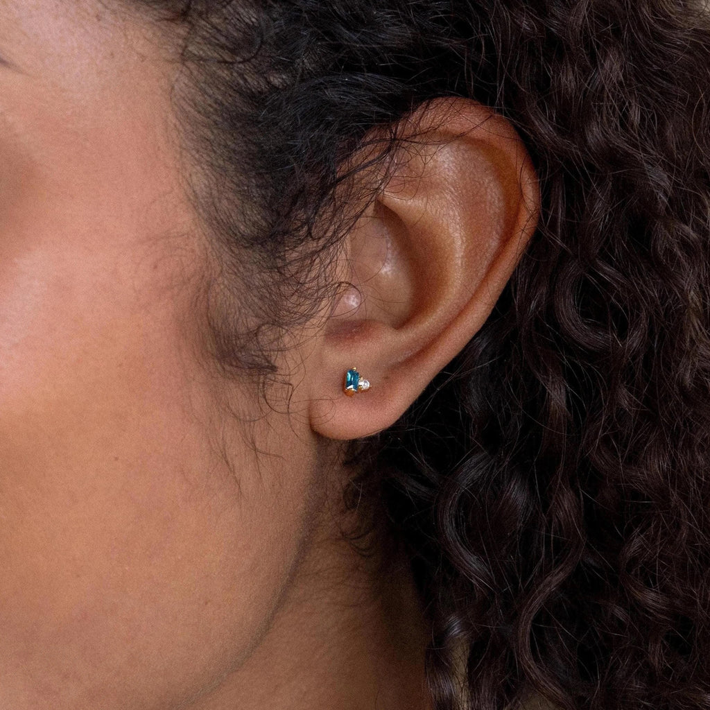 Close-up of an ear wearing the Toi et Moi Baguette Birthstone Studs, small gold earrings featuring a blue and a clear gem.