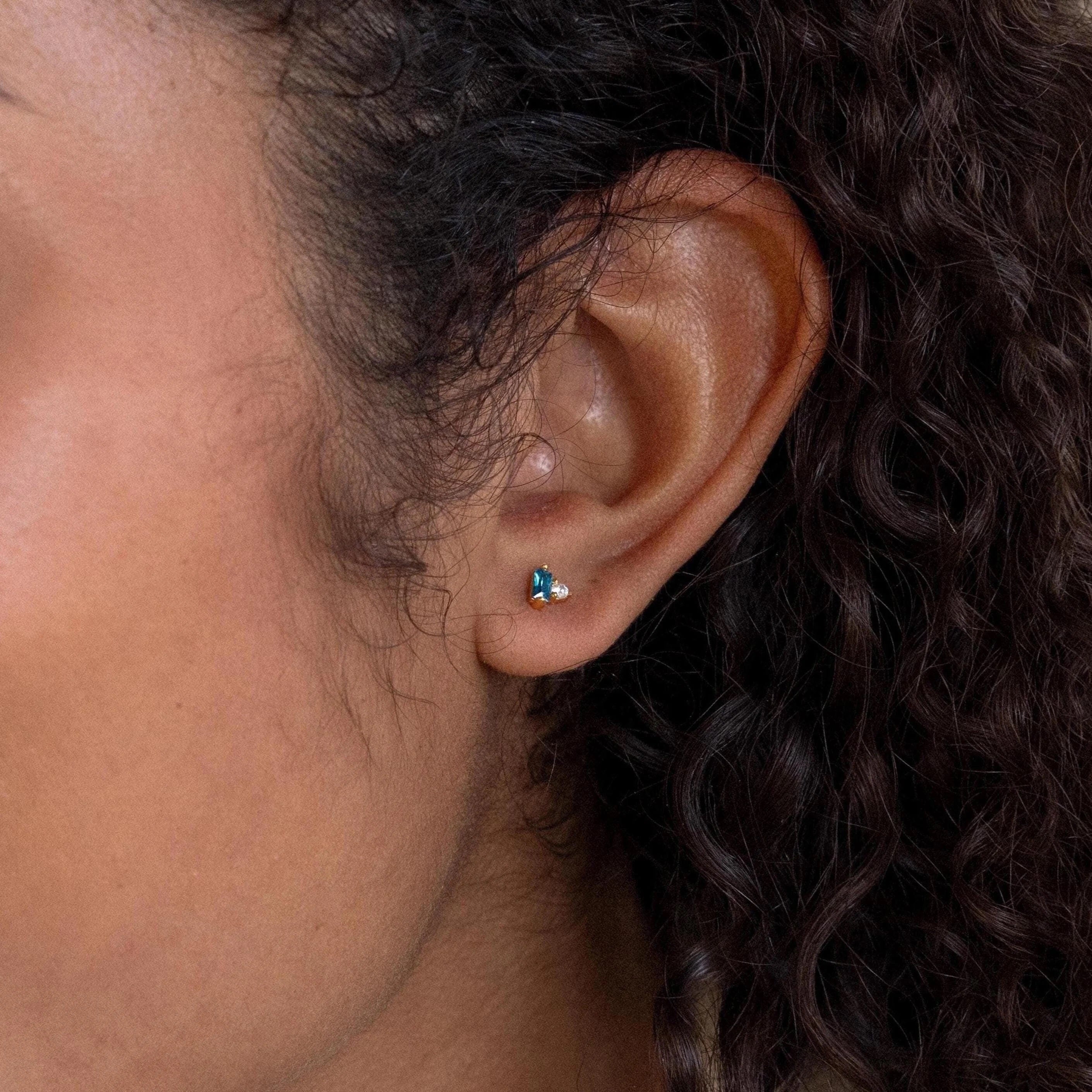 Close-up of an ear wearing the Toi et Moi Baguette Birthstone Studs, small gold earrings featuring a blue and a clear gem.