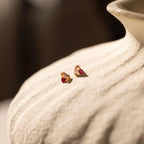 The Toi et Moi Baguette Birthstone Studs are small gold earrings with red gemstones, shown on a textured light ceramic surface.