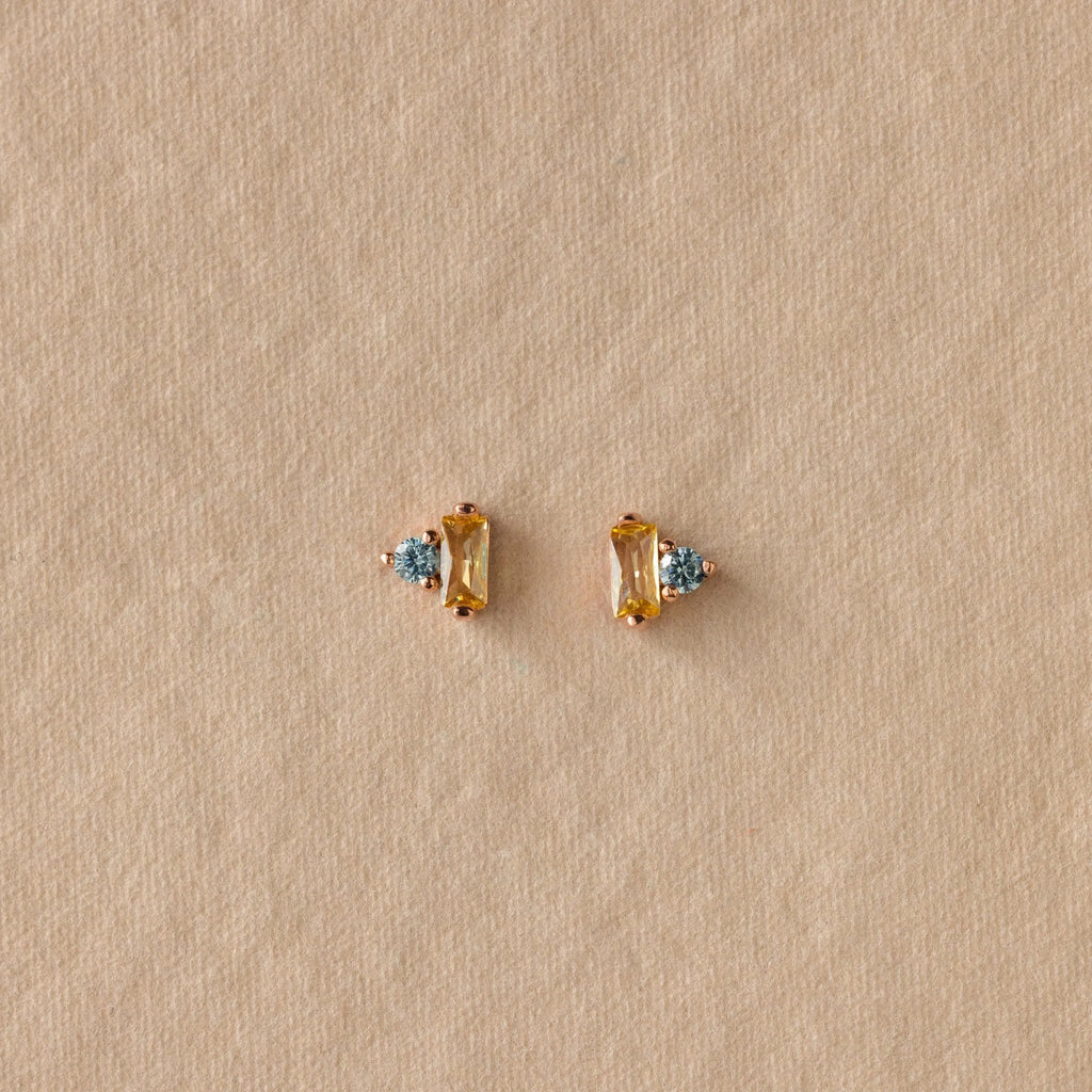 Toi et Moi Baguette Birthstone Studs in gold with blue gemstones are displayed on a textured beige background.