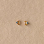 Toi et Moi Baguette Birthstone Studs in gold with blue gemstones are displayed on a textured beige background.