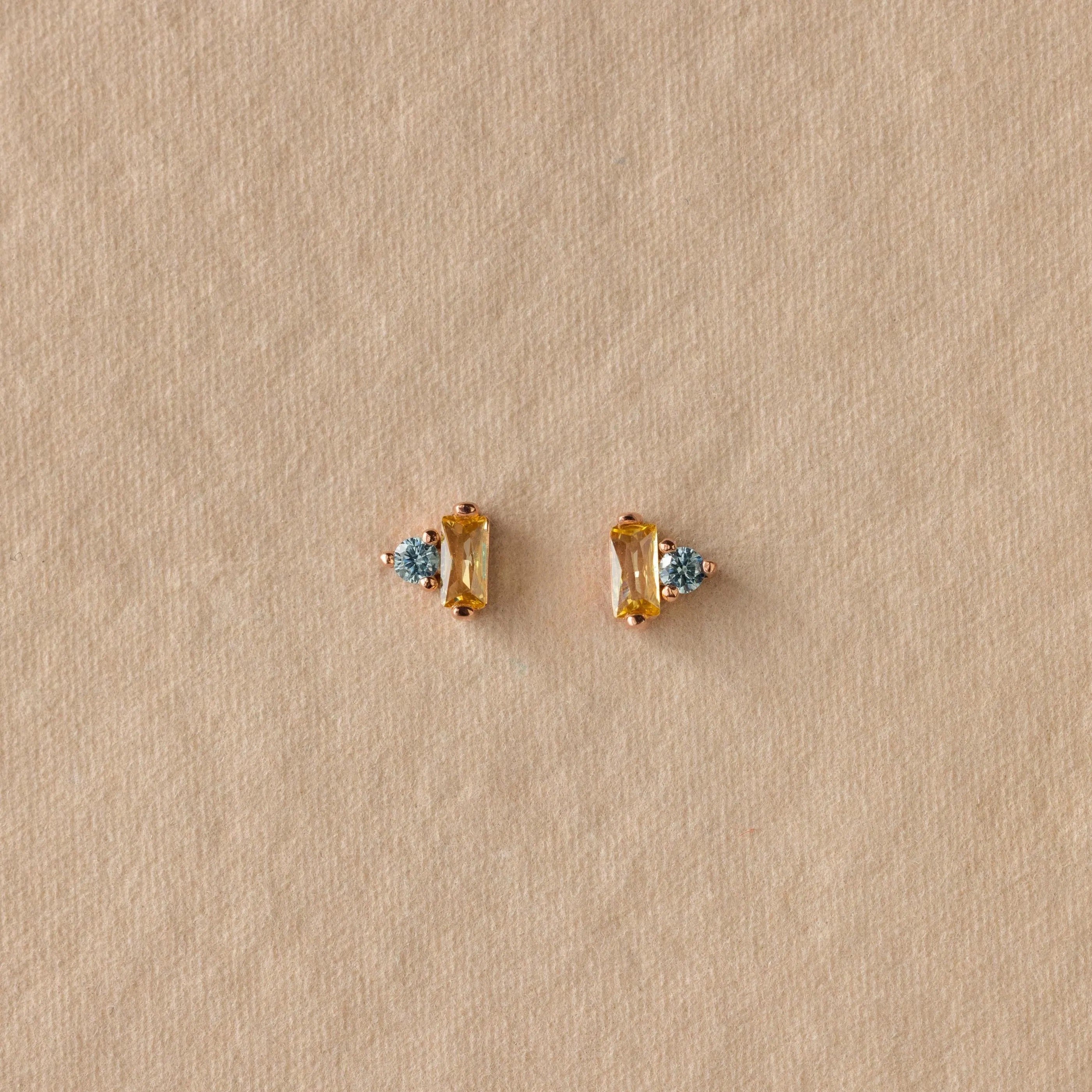 Toi et Moi Baguette Birthstone Studs in gold with blue gemstones are displayed on a textured beige background.
