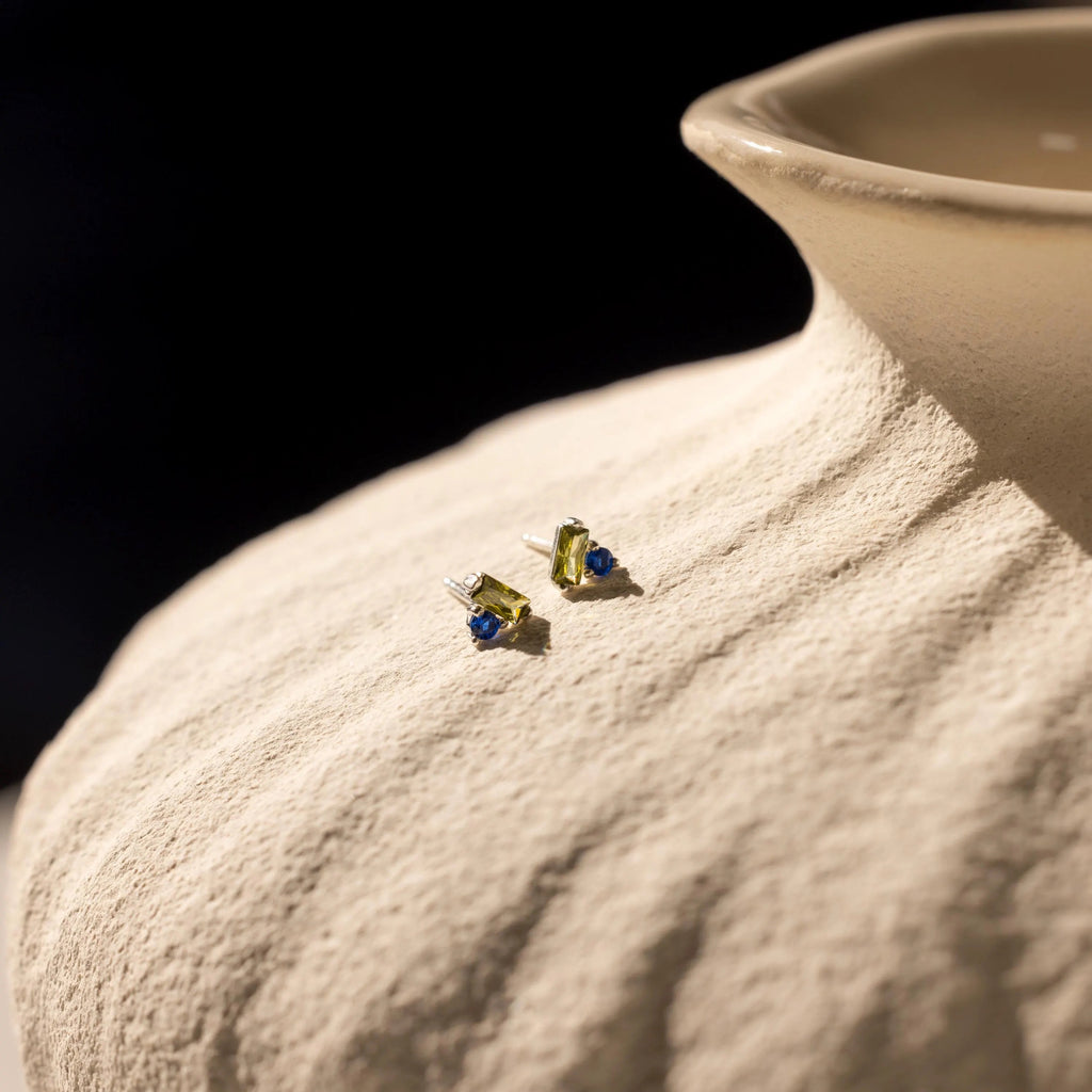 Toi et Moi Baguette Birthstone Studs featuring green and blue gemstones are displayed on a textured beige ceramic vase.