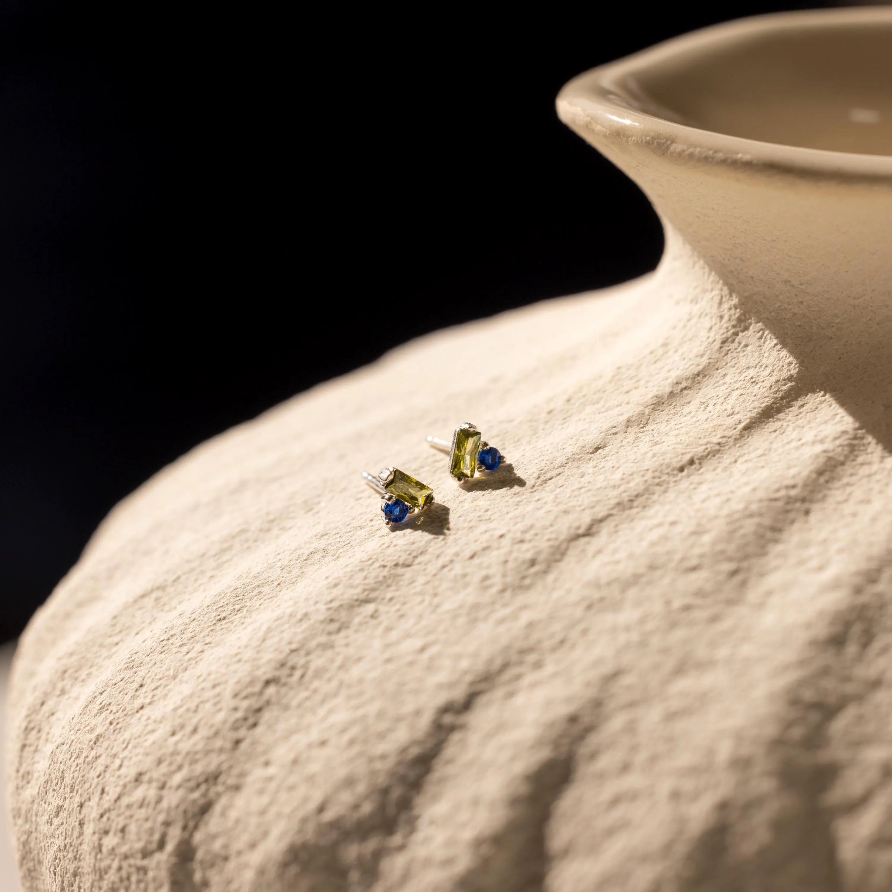 Toi et Moi Baguette Birthstone Studs featuring green and blue gemstones are displayed on a textured beige ceramic vase.