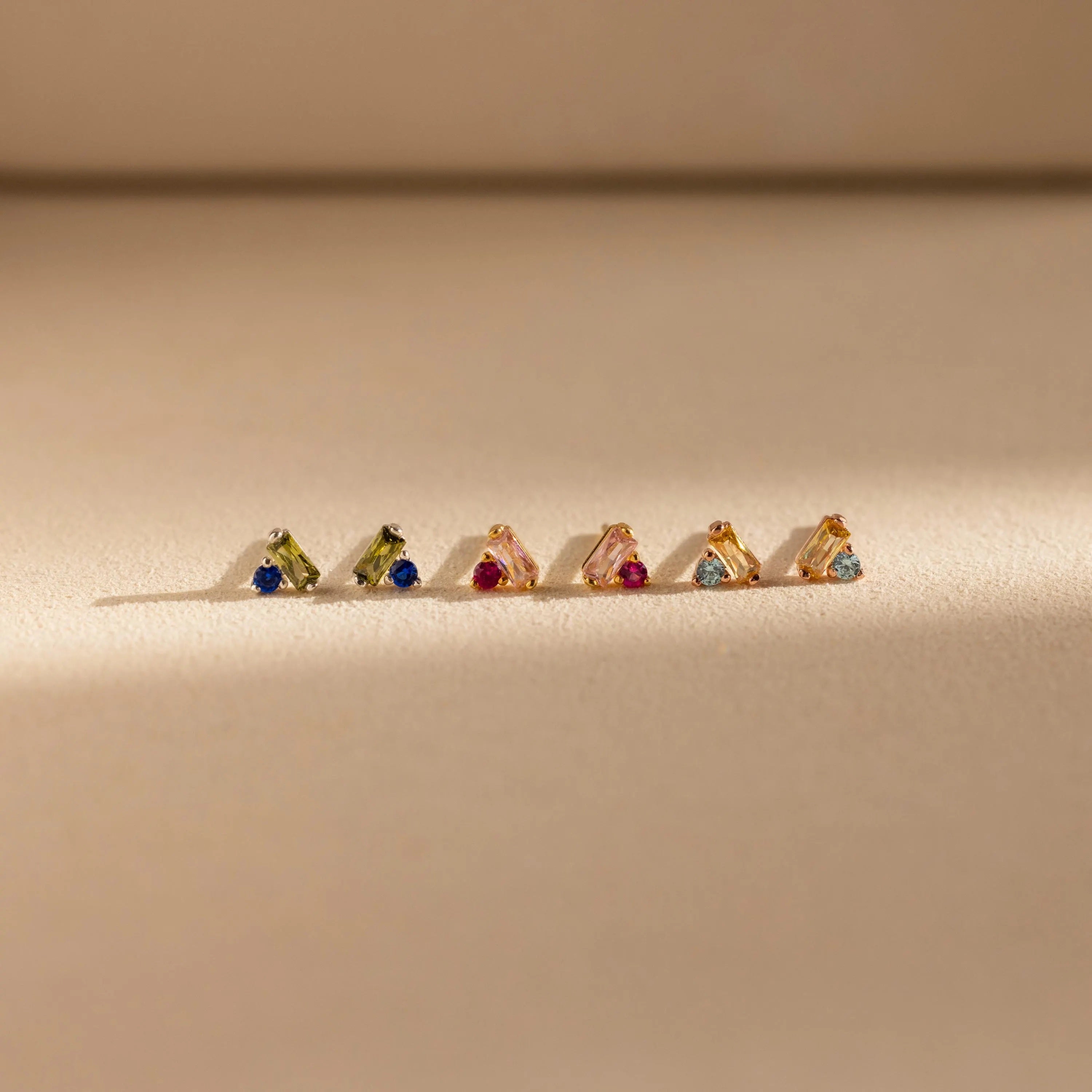 Six pairs of gold earrings, including Toi et Moi Baguette Birthstone Studs and other vibrant styles, are neatly arranged on a beige surface in soft light.