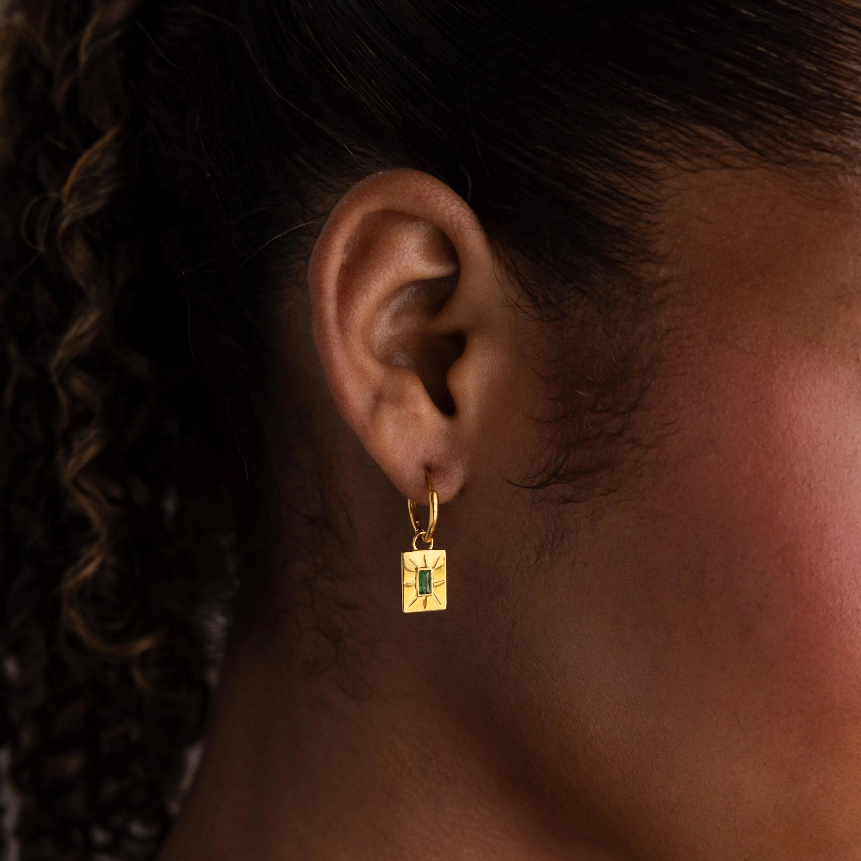 Close-up of a person wearing Birthstone Tag Huggies—gold rectangular earrings with a green gemstone, ideal for personalized jewelry lovers.